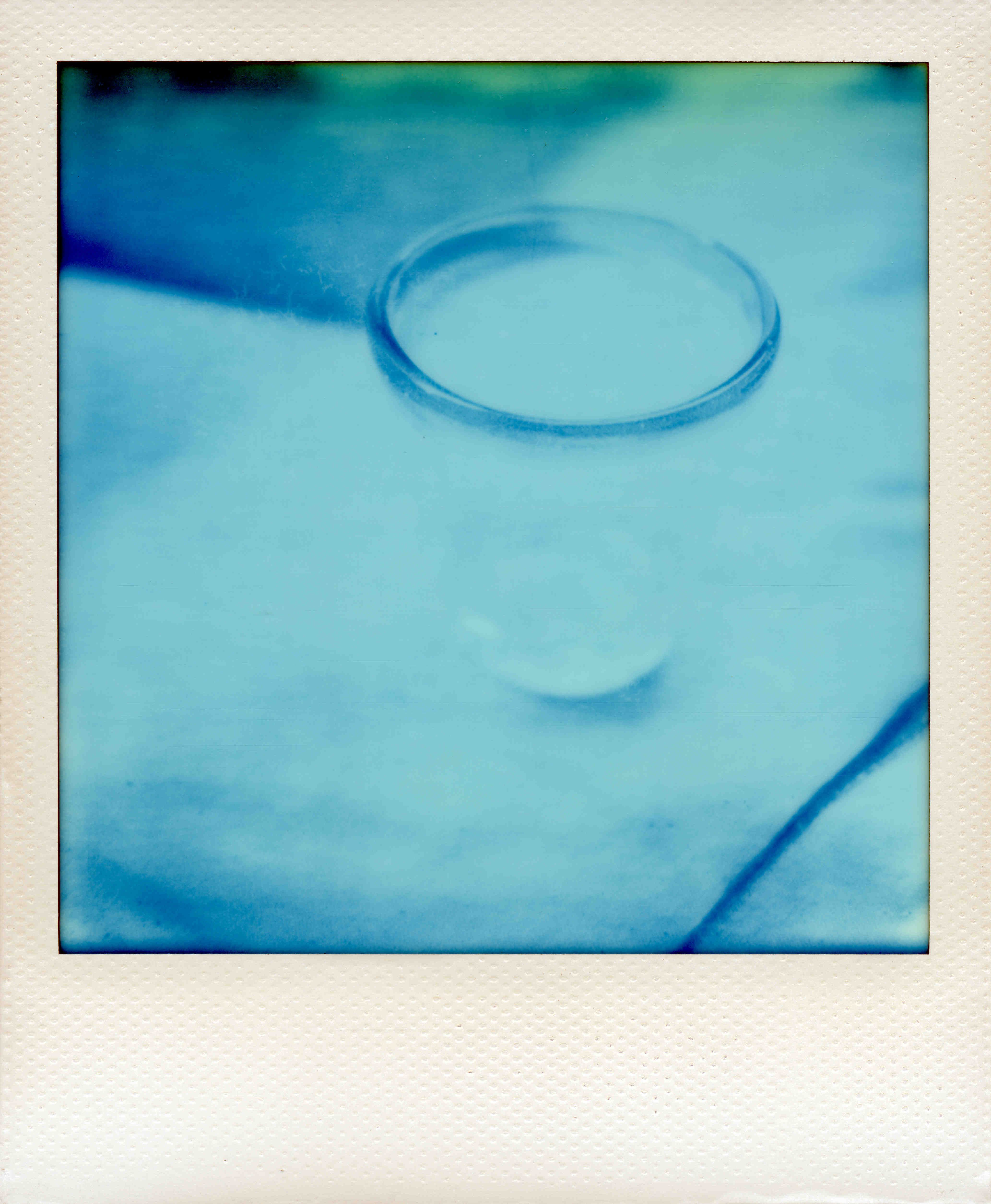 Ein Sofortbild mit weißem Bildrand, das Bild ist blau und zeigt ein Wasserglas mit dunklem Rand.