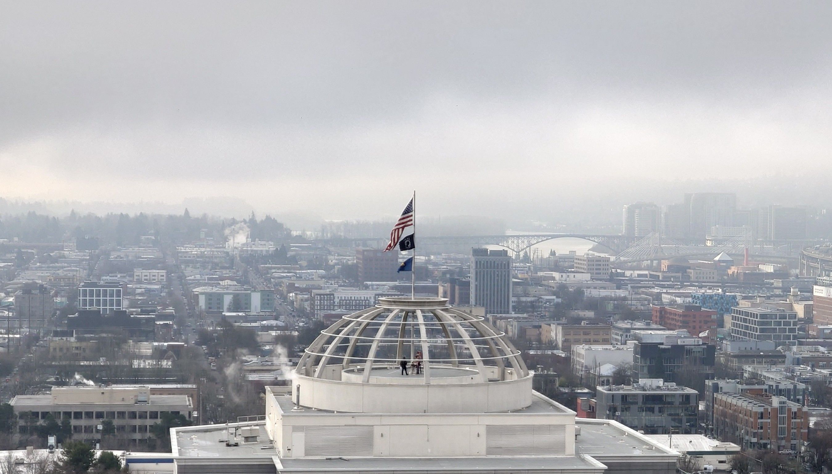 Flag of the "United States of America" after being winched up over a state building in Portland, Oregon. Two people standing inside the glassless dome are still present after raising it from whatever state it was previously in.

The day is misty and cloudy.