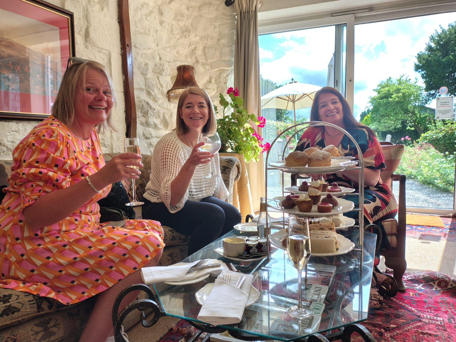 Three lassés sitting round a table , on the table is the typical British afternoon tea sandwich and cake stand, the lasses are raising glasses of  fizz .