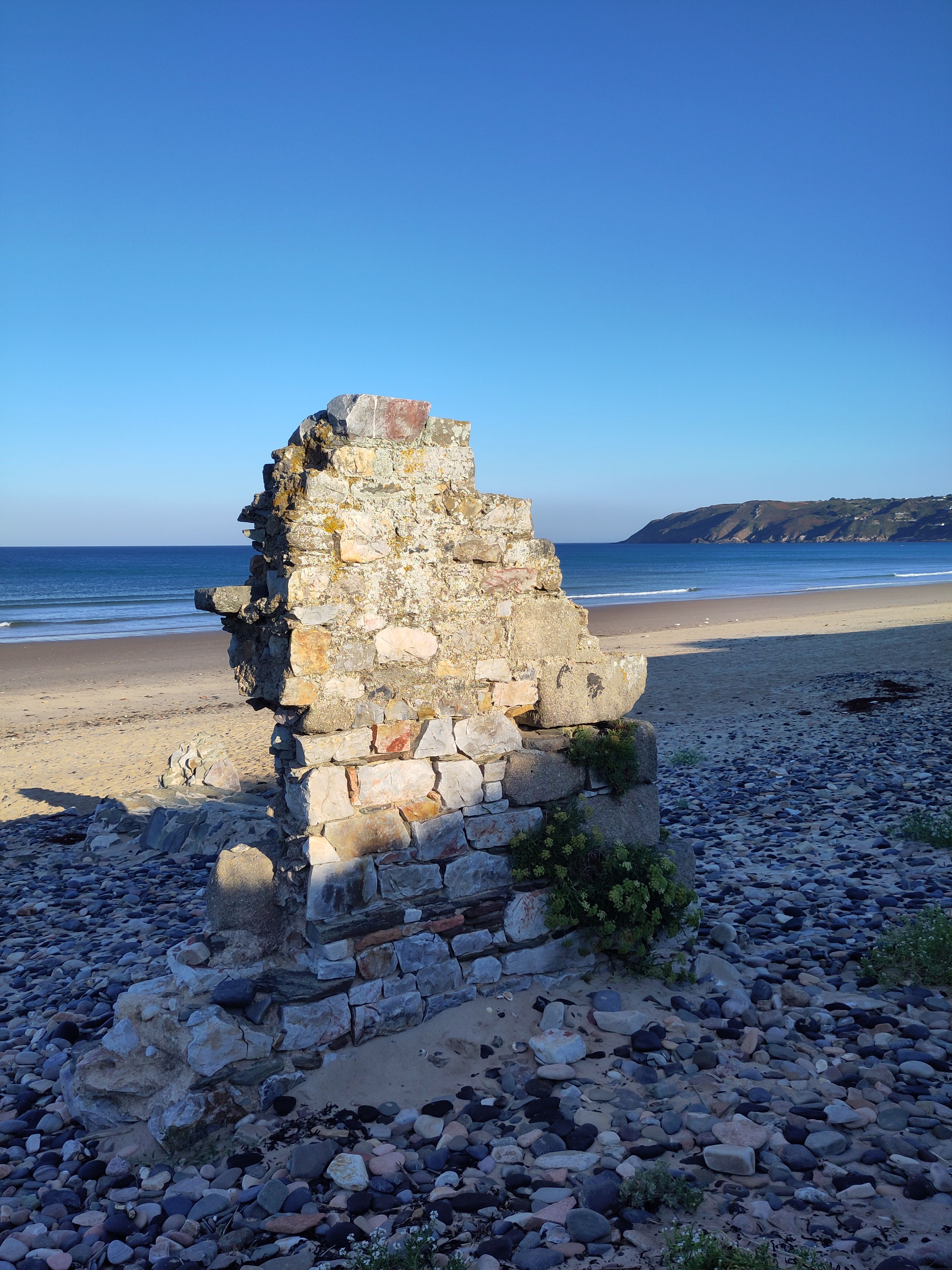 Ruin of stone building on beach with headland in background.