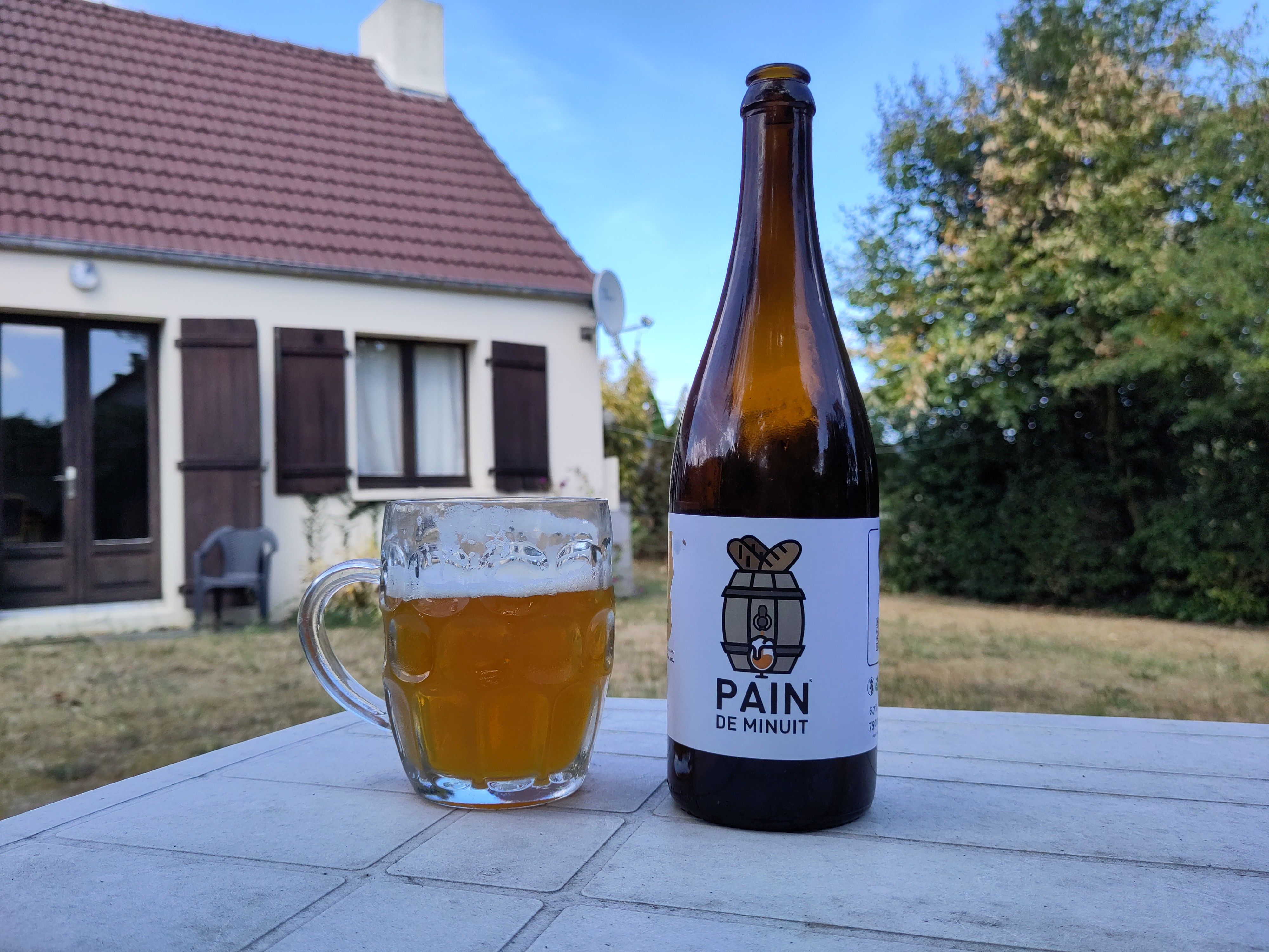 Pain De Minut bier bottle and beer mug on table with Norman traditional house in background.